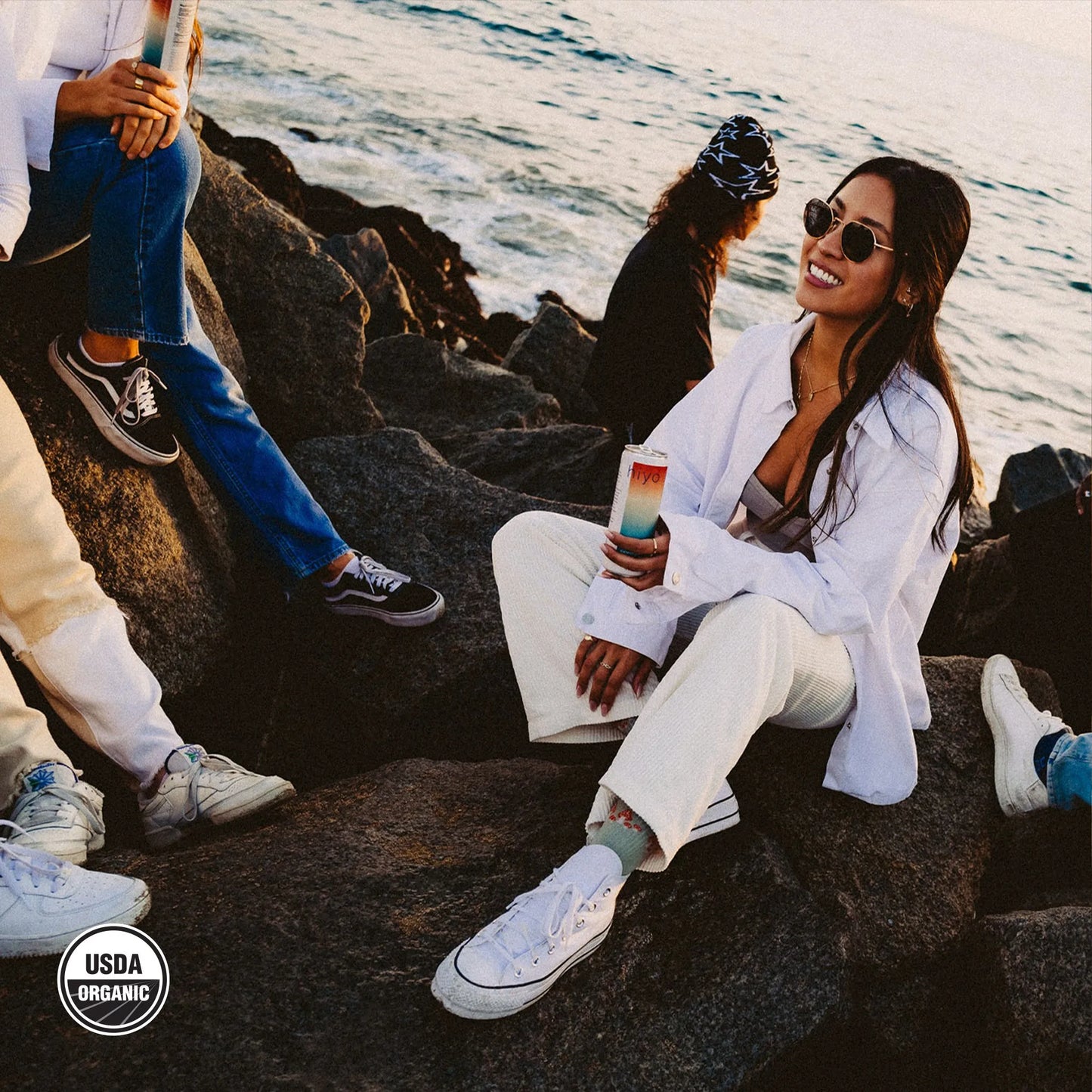 a woman hanging out with others on beach rocks while holding a can of hiyo watermelon lime