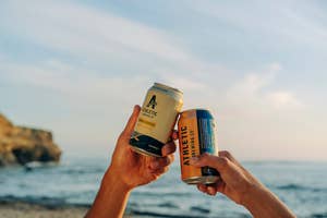 hands clinking two athletic brewing cans together in front of the ocean