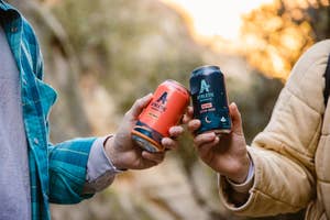 two athletic brewing co cans being clinked together by figures in jackets. the background is a lush hiking trail