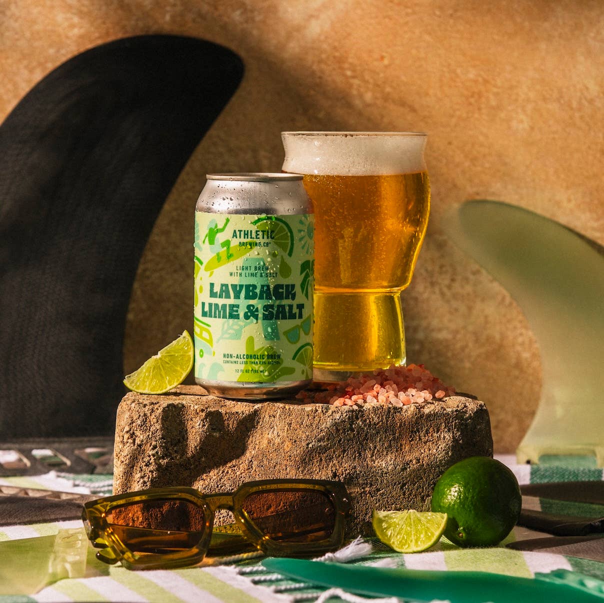 a can of layback lime & salt placed atop a rock with a glass full of the drink and a lime slice.  the rock is placed on a green outdoor blanket, surrounding the rock is a pair of sunglasses, a lime and another lime slice