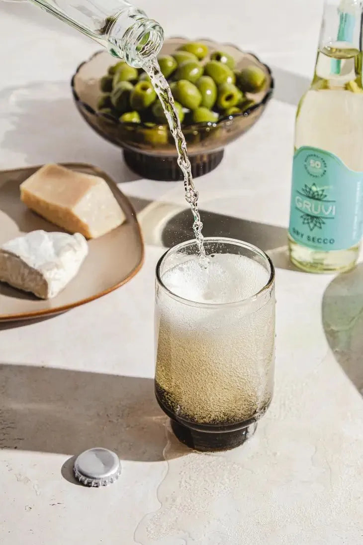 gruvi dry secco being poured into a glass next to a bottle, green olives and a charcuterie board